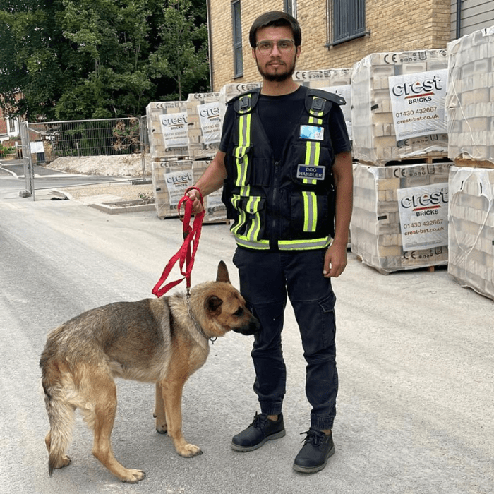 A security dog handler wearing a reflective vest stands with a German Shepherd on a construction site, showcasing professional Empty Property Security Services by 2015 Security Services Ltd.